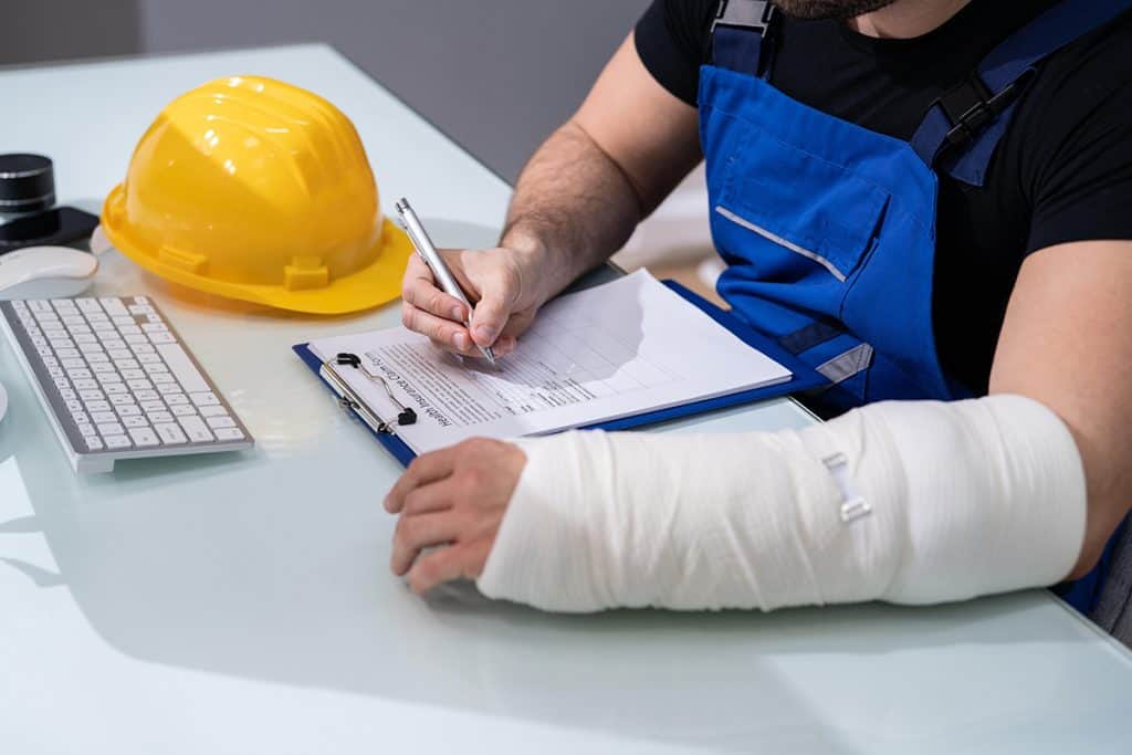 trabajador con mano escayolada sentado en una silla y escribiendo en un papel con la otra mano